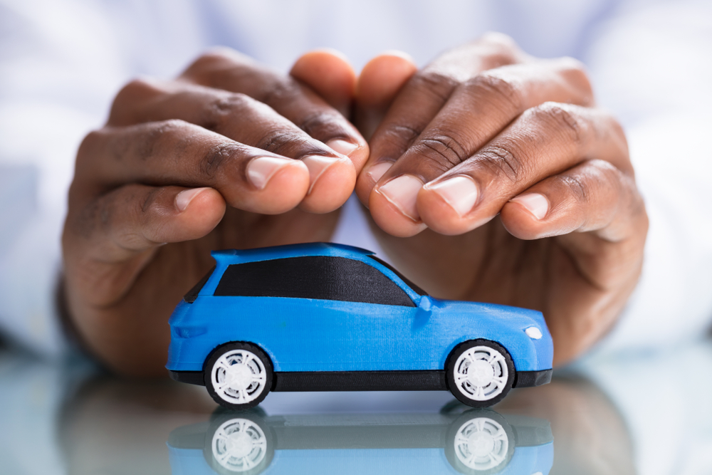 Shutterstock_1158955357 Businessman's Hand Protecting Blue Toy Car On The Reflective Desk