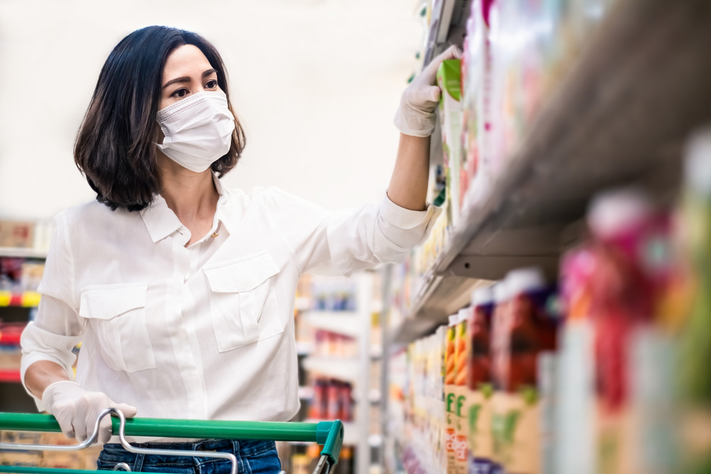 Shutterstock_1708963345 Asian woman wearing face mask and rubber glove push shopping cart in suppermarket departmentstore. Girl choosing, looking grocery things to buy at shelf during coronavirus crisis or covid19 outbreak.