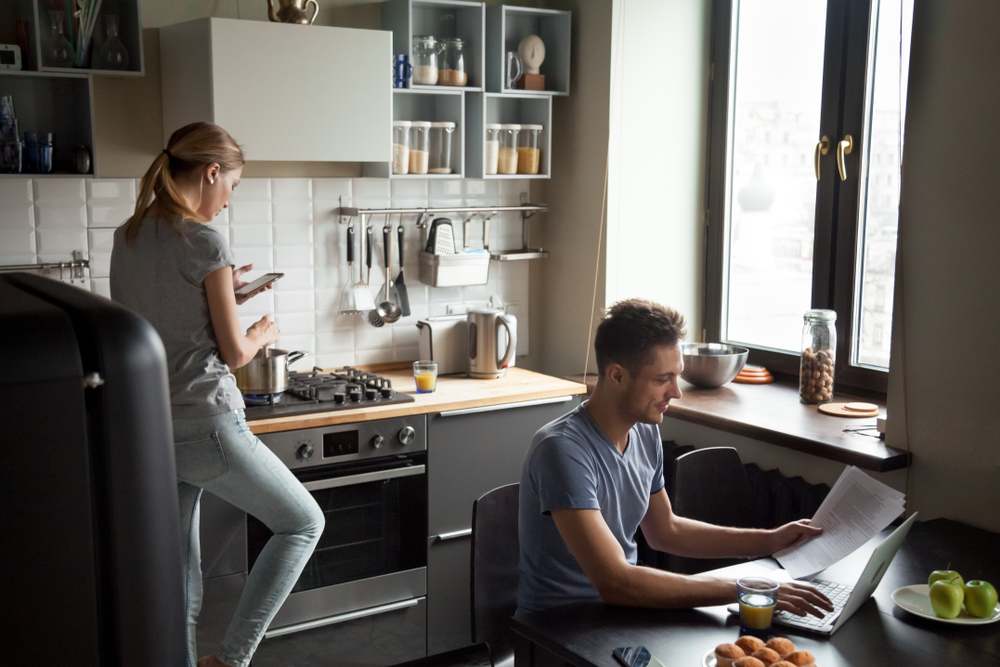 Shutterstock_1156208539 Young couple using laptop and smartphone in the kitchen, smiling man working online at home while his wife cooking with phone apps, family morning lifestyle with gadgets or devices addiction concept