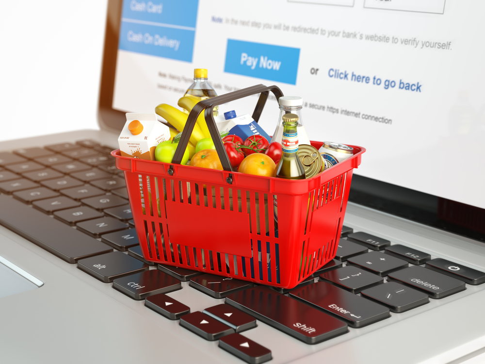 Shutterstock_581283208 Shopping basket with variety of grocery products ion laptop keyboard. E-commerce concept 3d illustration